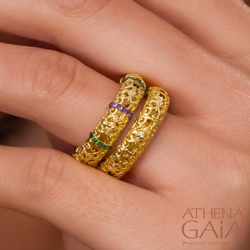 Two 18k gold rings with intricate embroidery and multicolored stones on a person's fingers.