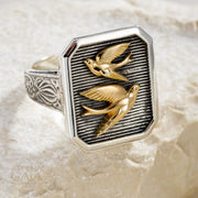 Gerochristo Silver ring with two gold swallow birds on a textured beige background