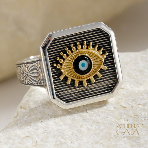 925 Sterling Silver square ring with an 18k Gold evil eye design on a stone surface, branded 'Athena Gaia'.