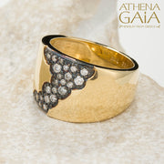 A wide band 18k gold ring with a pattern of inset black and white diamonds along the top side, displayed on a stone surface.