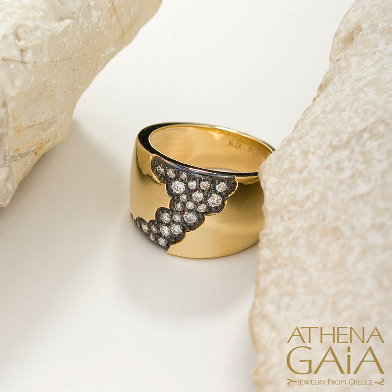 A wide band 18k gold ring with a pattern of inset black and white diamonds along the top side, displayed on a stone surface.