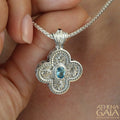 Intricate silver cross pendant with ornate filigree and central blue gemstone held on a silver chain.