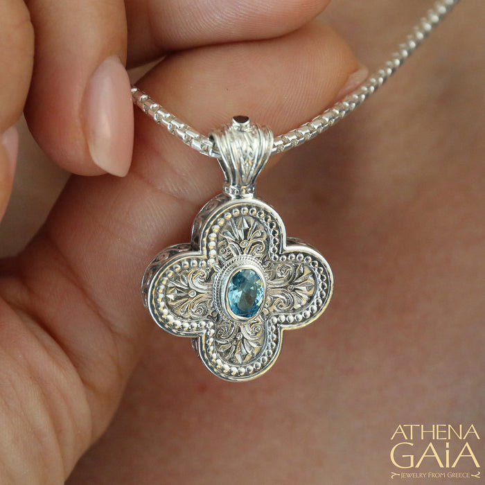 Intricate silver cross pendant with ornate filigree and central blue gemstone held on a silver chain.