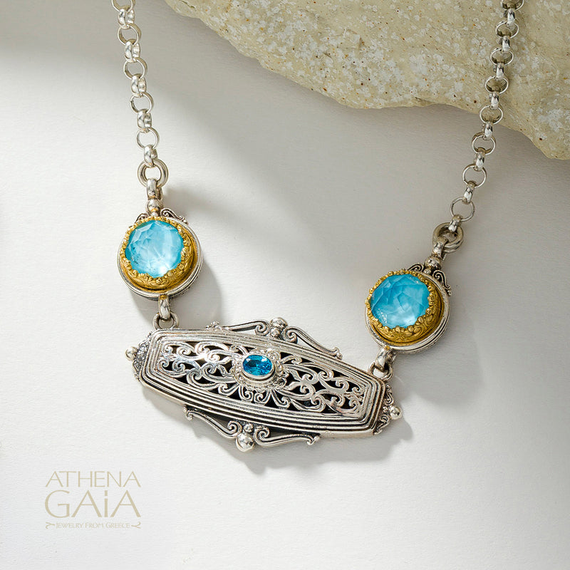 An ornate silver filigree pendant necklace with two round light blue gemstones in gold bezels and a small blue stone in the center, displayed on a light surface with a stone prop and the Athena Gaia logo in the corner.
