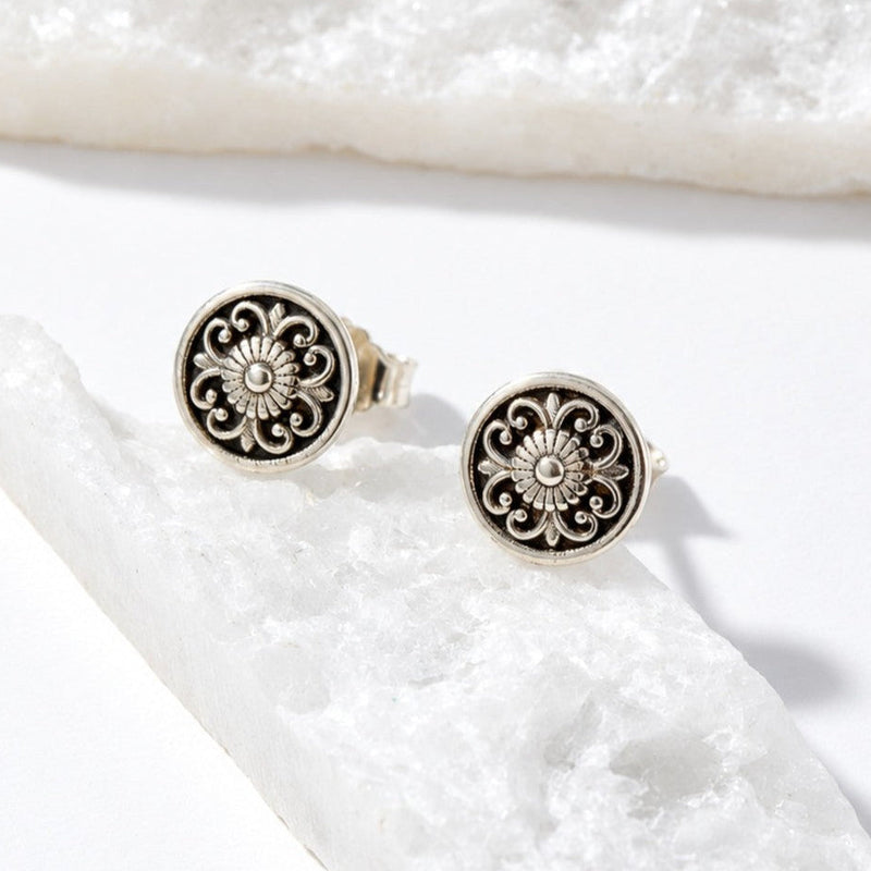 A pair of round silver-toned stud earrings with an ornate filigree floral design, displayed on a white textured crystal against a clean white background.