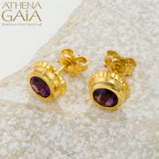 Gold stud earrings with deep purple gemstones, elegantly displayed on a light textured stone.