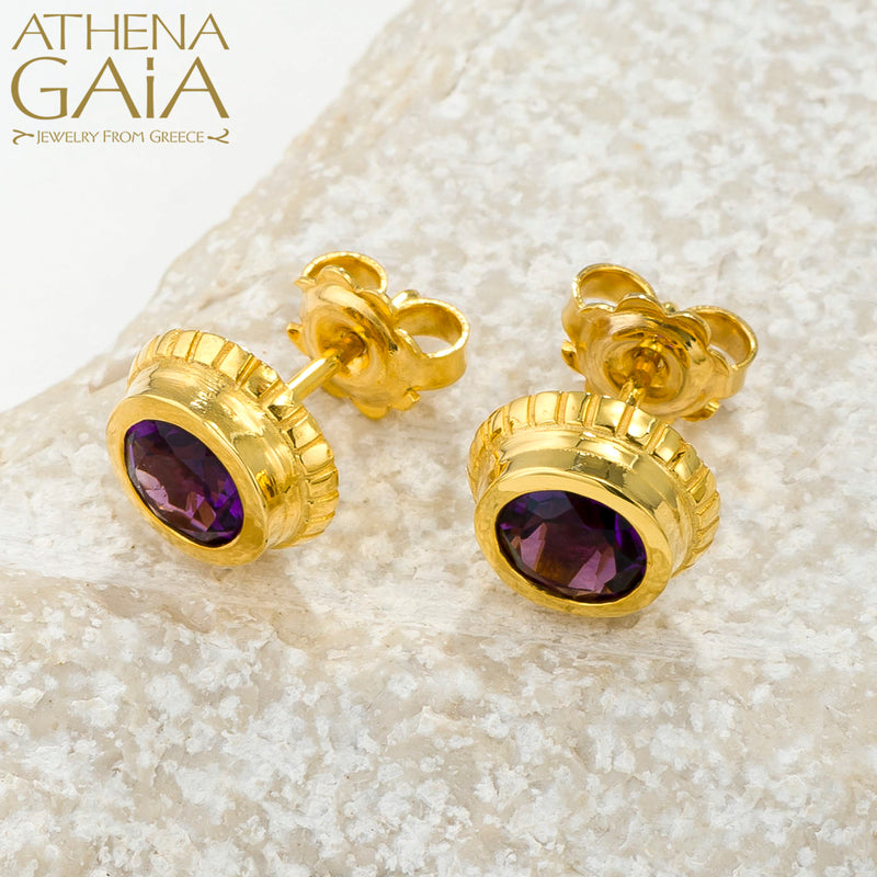 Gold stud earrings with deep purple gemstones, elegantly displayed on a light textured stone.