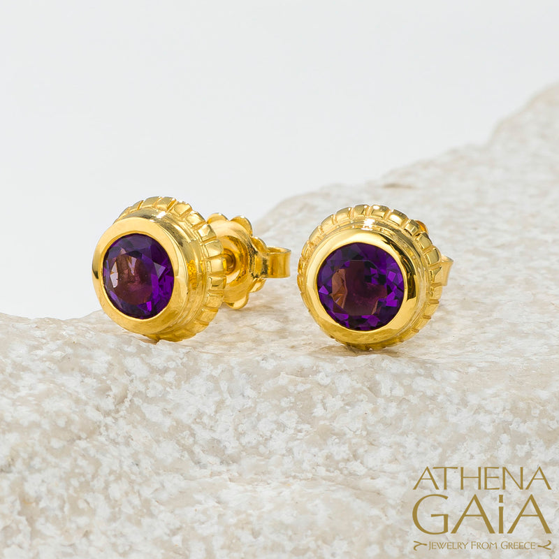 Gold stud earrings with deep purple gemstones, elegantly displayed on a light textured stone.
