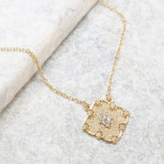 Delicate gold necklace featuring a textured square pendant adorned with small sparkling white stones at its center.