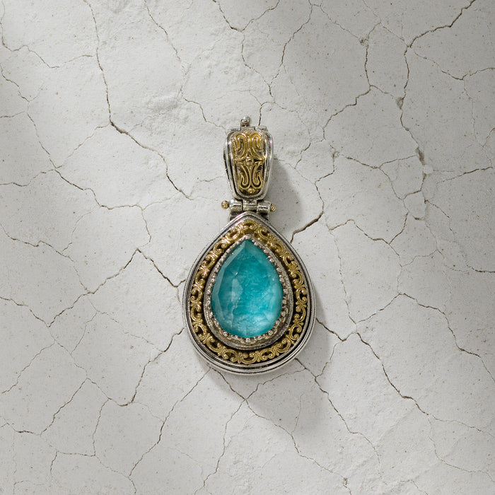 Teardrop shaped pendant with a bright blue stone framed by ornate silver and gold detailing, displayed on a cracked, textured surface.