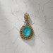 Teardrop shaped pendant with a bright blue stone framed by ornate silver and gold detailing, displayed on a cracked, textured surface.