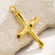 Gold cross pendant with green gemstones on a textured stone surface, branded 'Athena Gaia'.