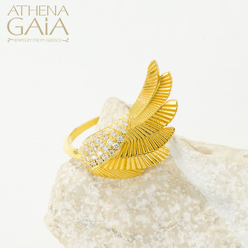 A gold-colored ring with a large wing-like design adorned with small white stones, displayed on a white surface.
