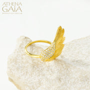 A gold-colored ring with a large wing-like design adorned with small white stones, displayed on a white surface.