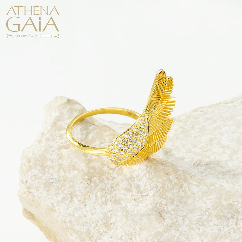 A gold-colored ring with a large wing-like design adorned with small white stones, displayed on a white surface.