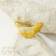 A gold-colored ring with a large wing-like design adorned with small white stones, displayed on a white surface.