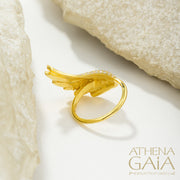 A gold-colored ring with a large wing-like design adorned with small white stones, displayed on a white surface.