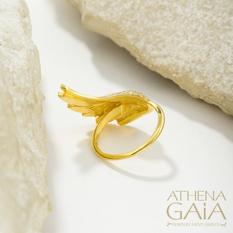 A gold-colored ring with a large wing-like design adorned with small white stones, displayed on a white surface.