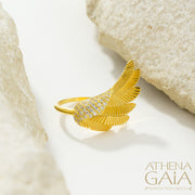 A gold-colored ring with a large wing-like design adorned with small white stones, displayed on a white surface.