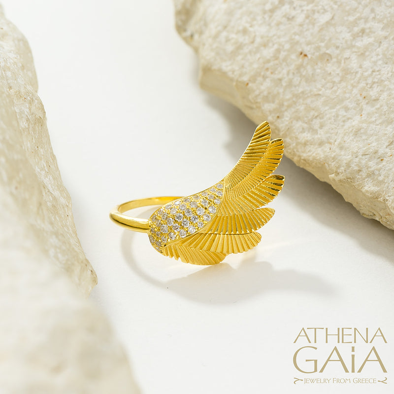 A gold-colored ring with a large wing-like design adorned with small white stones, displayed on a white surface.