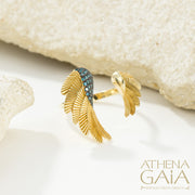 A gold-colored ring with leaf-like designs and blue gemstones placed on a white surface, with the brand name 'ATHENA GAIA' visible.