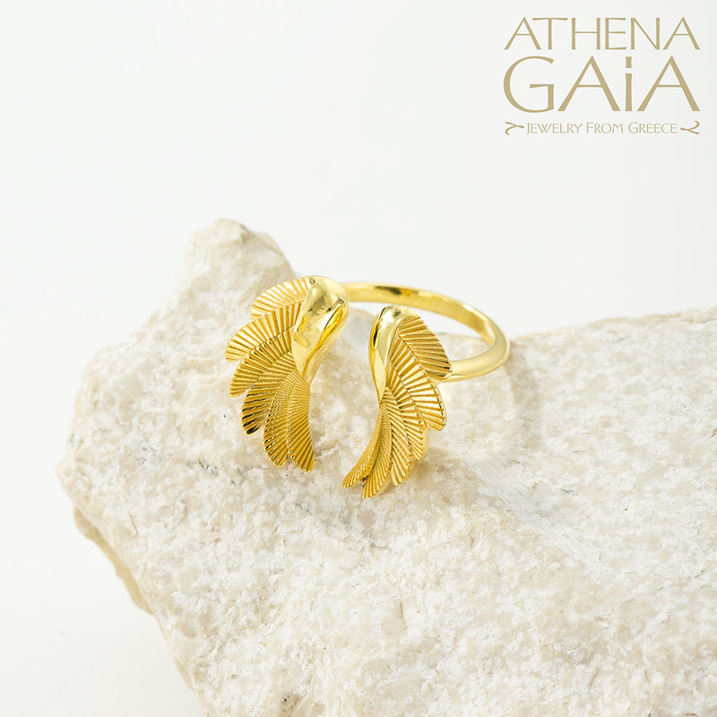 A gold-colored ring with wing-like designs displayed on a textured white surface, with the brand name 'ATHENA GAIA' visible.