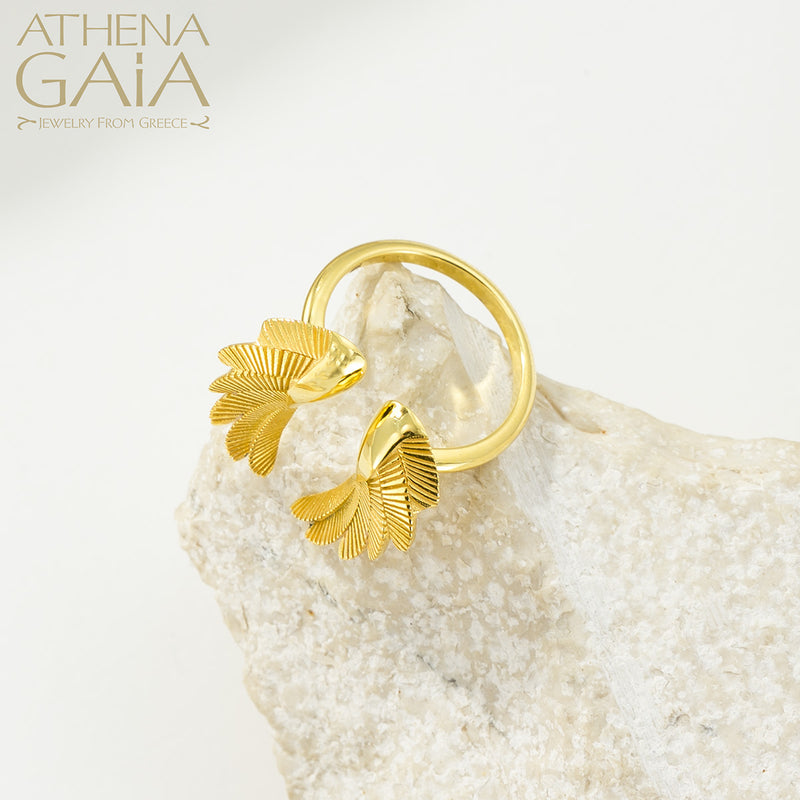 A gold-colored ring with wing-like designs displayed on a textured white surface, with the brand name 'ATHENA GAIA' visible.