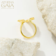 A gold-colored ring with wing-like designs displayed on a textured white surface, with the brand name 'ATHENA GAIA' visible.