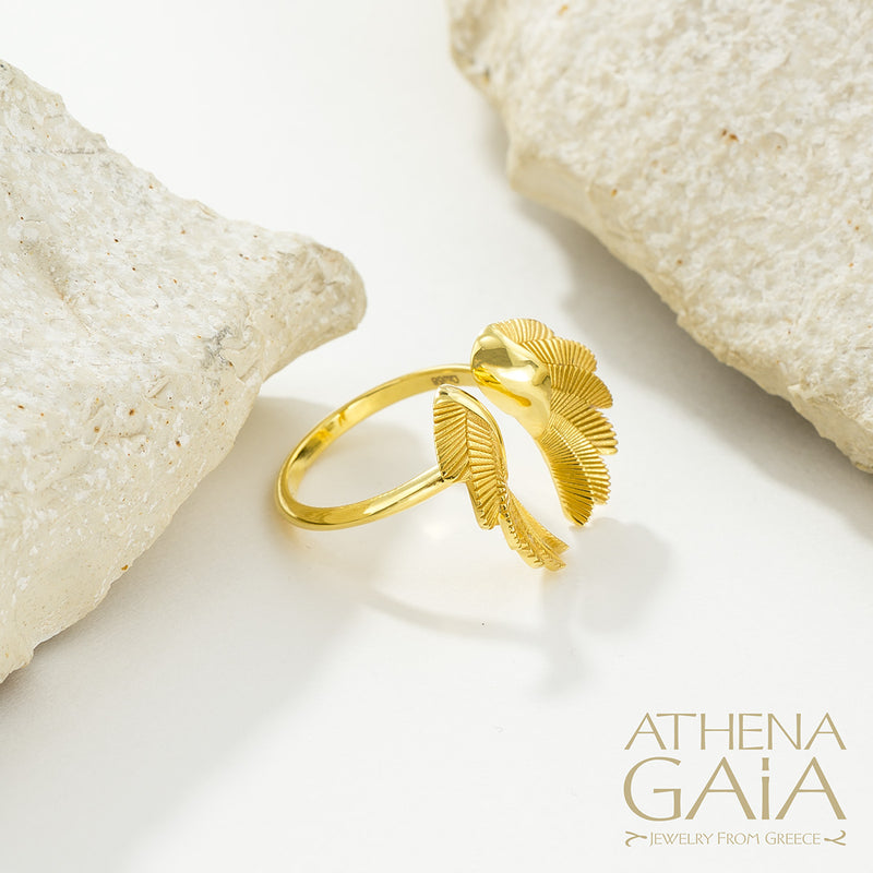 A gold-colored ring with wing-like designs displayed on a textured white surface, with the brand name 'ATHENA GAIA' visible.