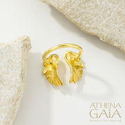 A gold-colored ring with winged motifs, displayed on a textured surface with the brand name 'ATHENA GAIA' visible.