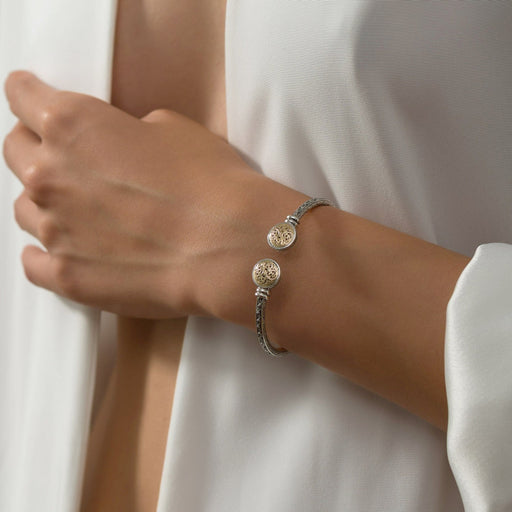 A close-up of a person's wrist wearing a silver-toned chain bracelet with two round, textured gold-toned charms, styled with a white draped top.