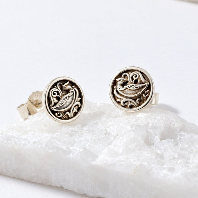 A pair of round silver stud earrings with an embossed bird design, displayed on a white textured stone slab against a clean white background.