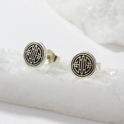 A pair of round silver-toned stud earrings with a black geometric monogram design, displayed on a white textured stone slab.