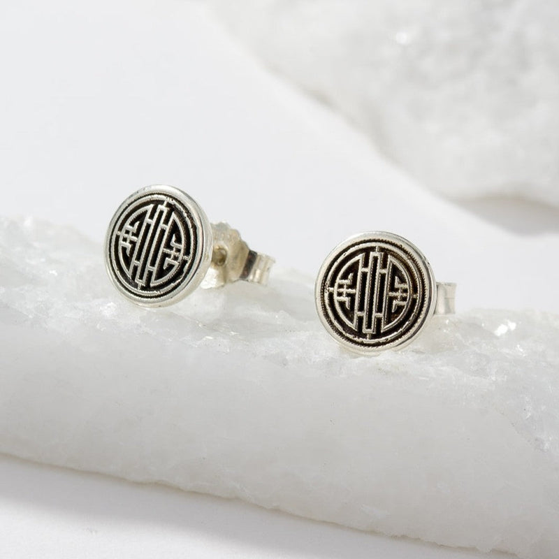 A pair of round silver-toned stud earrings with a black geometric monogram design, displayed on a white textured stone slab.