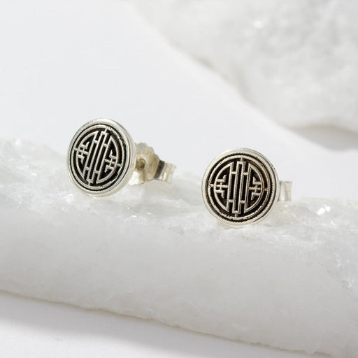A pair of round silver-toned stud earrings with a black geometric monogram design, displayed on a white textured stone slab.