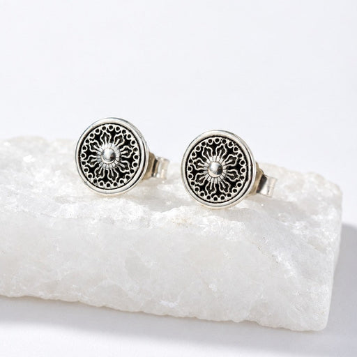 A pair of round silver-toned stud earrings with intricate filigree cutouts and a central sunburst design, displayed on a white crystal block against a white background.
