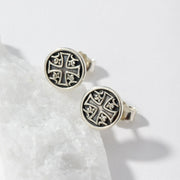 A pair of round silver-toned stud earrings with an engraved cross motif, displayed on a white textured stone slab against a clean white background.