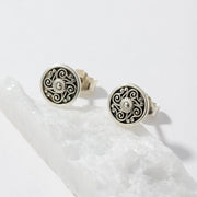 A pair of round silver-toned earrings with black filigree scrollwork and a small clear stone in the center, displayed on a white crystal against a white background.
