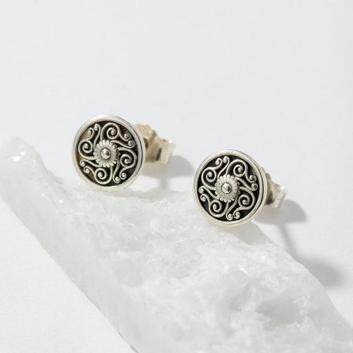 A pair of round silver-toned earrings with black filigree scrollwork and a small clear stone in the center, displayed on a white crystal against a white background.