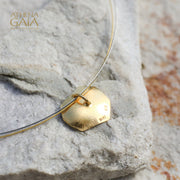 A gold-tone pendant necklace with a textured, organic-shaped tag-like pendant resting on a light gray stone, with a fine silver-tone wire running behind it. 