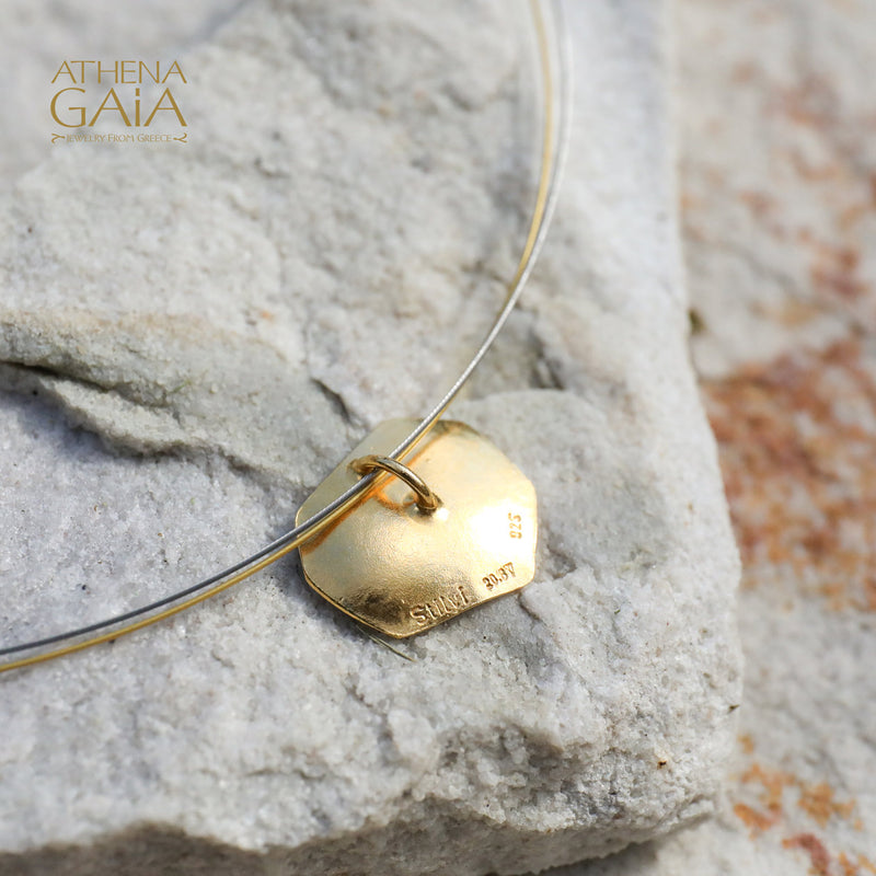 A gold-tone pendant necklace with a textured, organic-shaped tag-like pendant resting on a light gray stone, with a fine silver-tone wire running behind it. 