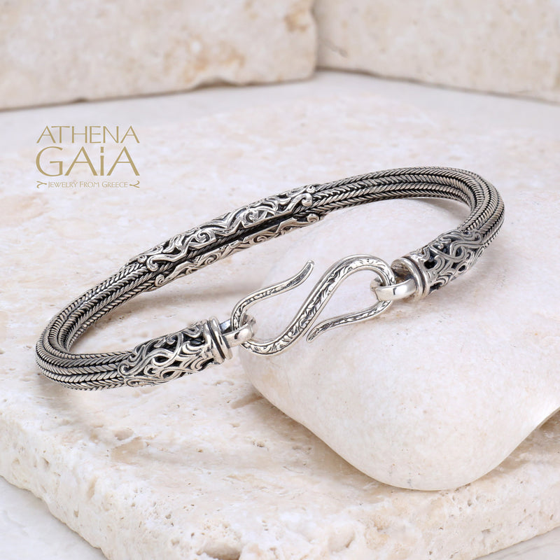 Intricate silver bracelet with detailed woven band and hook clasp, displayed on stone.