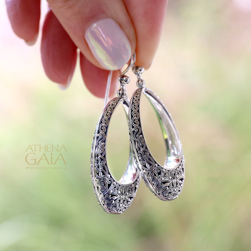 Hand holding ornate silver teardrop earrings with detailed filigree design and polished finish.