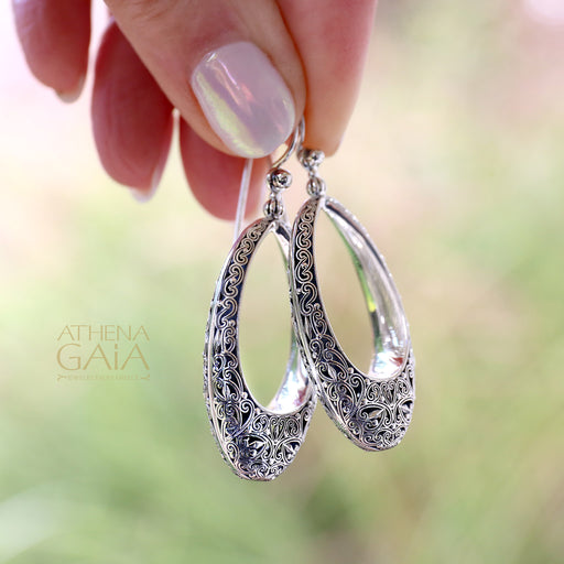 Hand holding ornate silver teardrop earrings with detailed filigree design and polished finish.