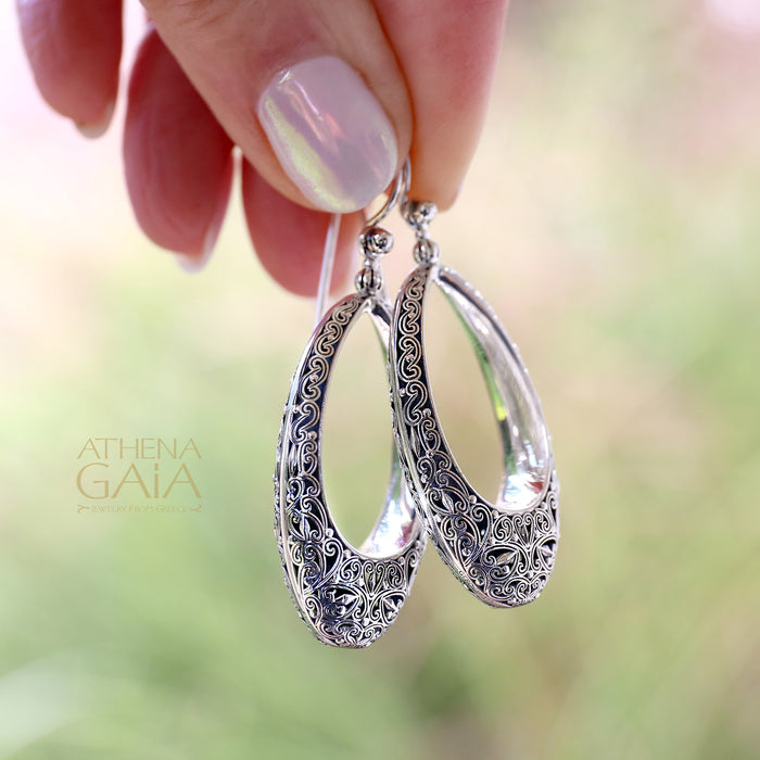 Hand holding ornate silver teardrop earrings with detailed filigree design and polished finish.