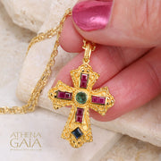 Gold cross pendant with ruby, emerald, and sapphire stones held by hand, on a gold chain necklace.