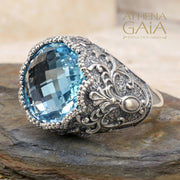 A sterling silver Byzantine filigree ring with a large blue topaz stone, set in a detailed filigree design on a rocky surface.
