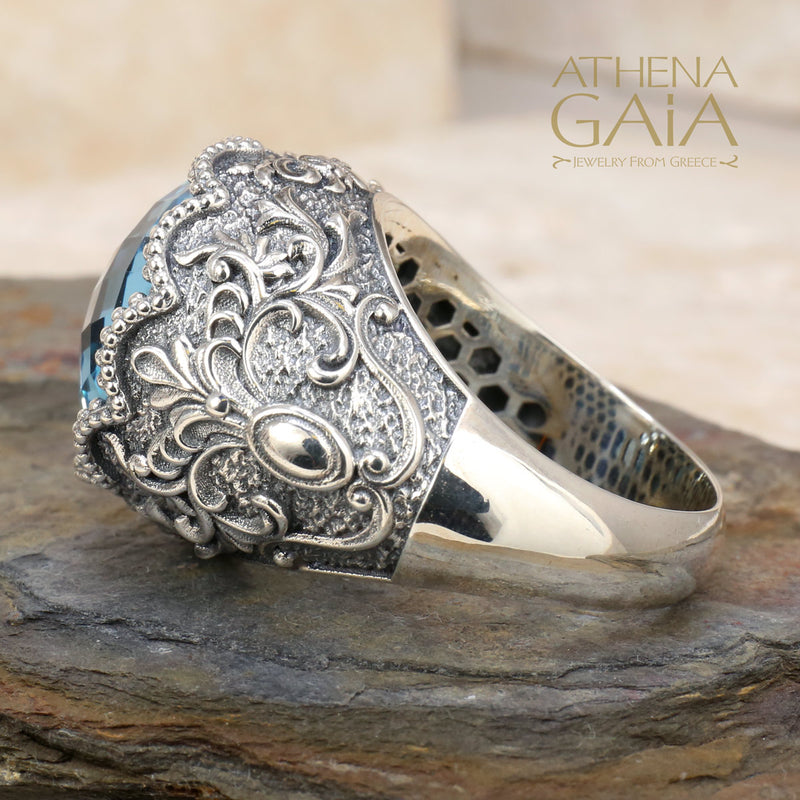 A sterling silver Byzantine filigree ring with a large blue topaz stone, set in a detailed filigree design on a rocky surface.