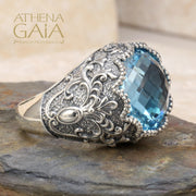 A sterling silver Byzantine filigree ring with a large blue topaz stone, set in a detailed filigree design on a rocky surface.