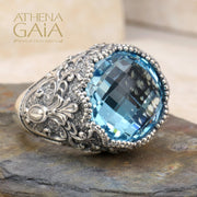 A sterling silver Byzantine filigree ring with a large blue topaz stone, set in a detailed filigree design on a rocky surface.